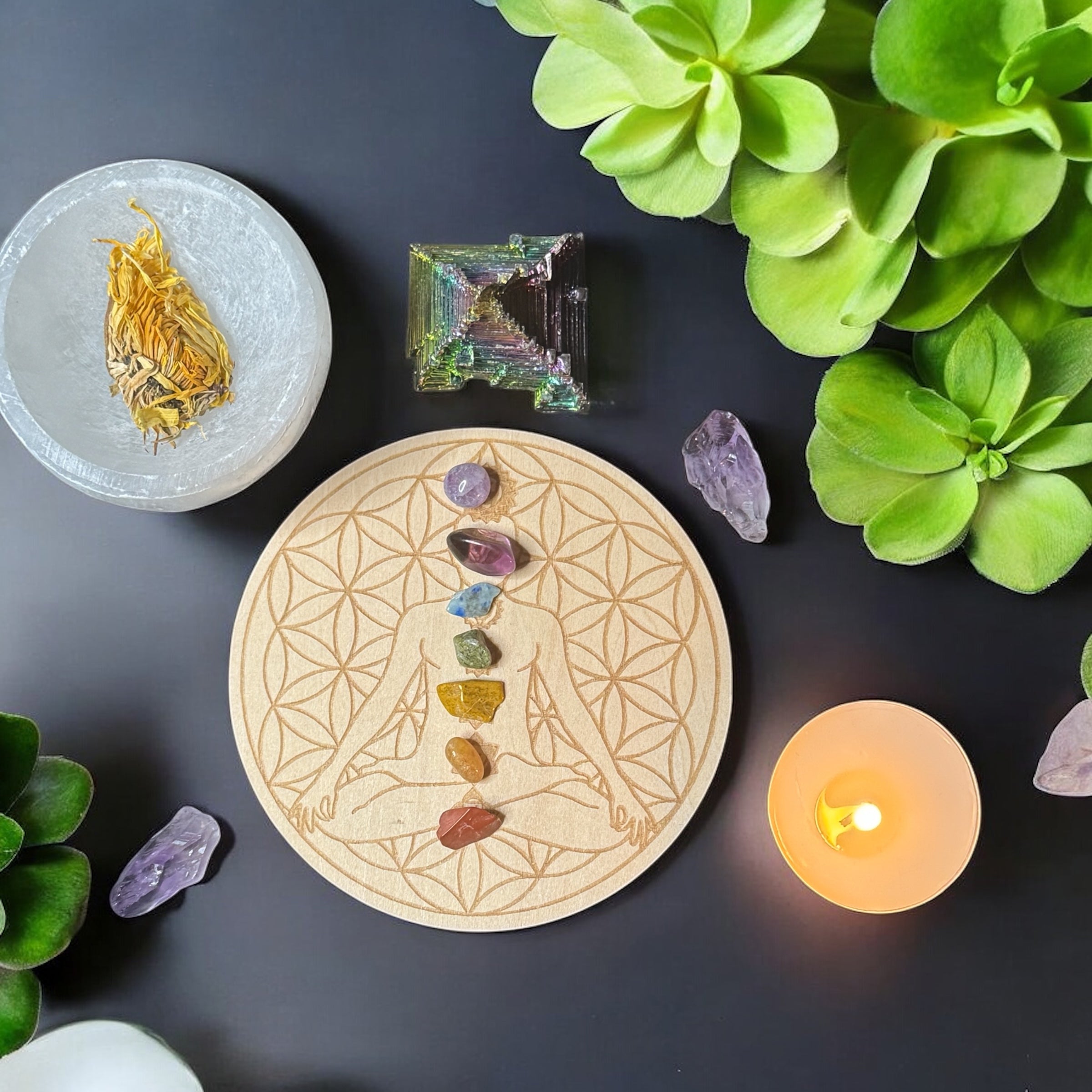 chakra colored crystals in a column sitting on a wooden circular board with a lit candle and a bowl of herbs 