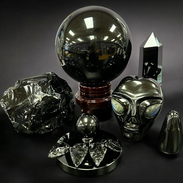 Black obsidian crystal tools including palm stones, towers, jewelry, and raw chunks displayed on natural wood with spiritual decor.